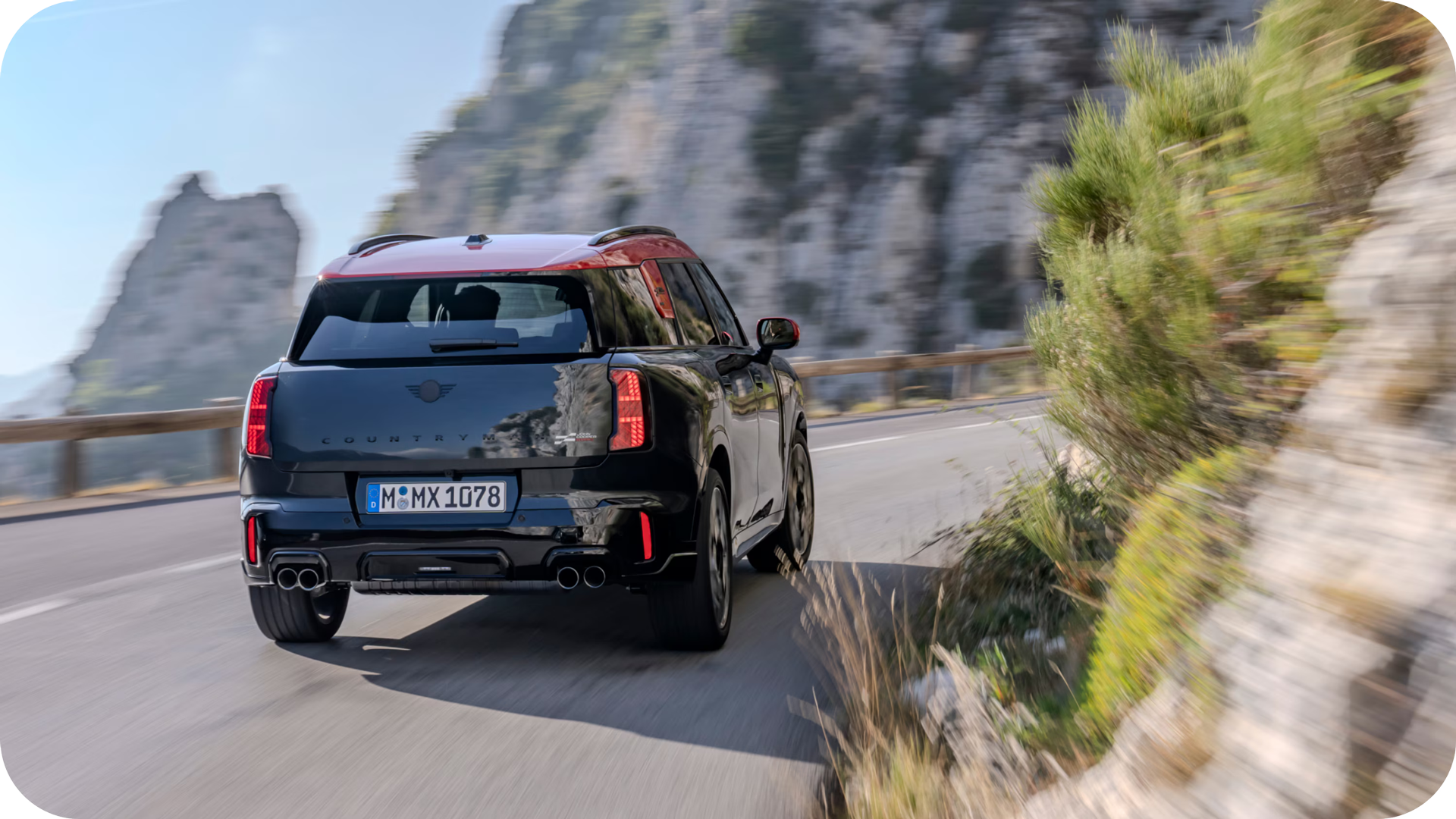 Rear-view of a 2025 MINI JCW Countryman ALL4 turning around a bend on a road with cliffs in the distance. 
